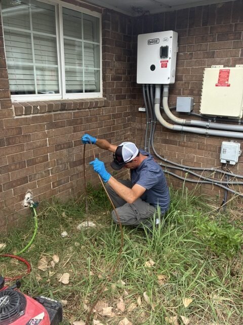 Plumber wearing blue gloves feeding a drain-cleaning line into an outdoor access point beside a brick building.